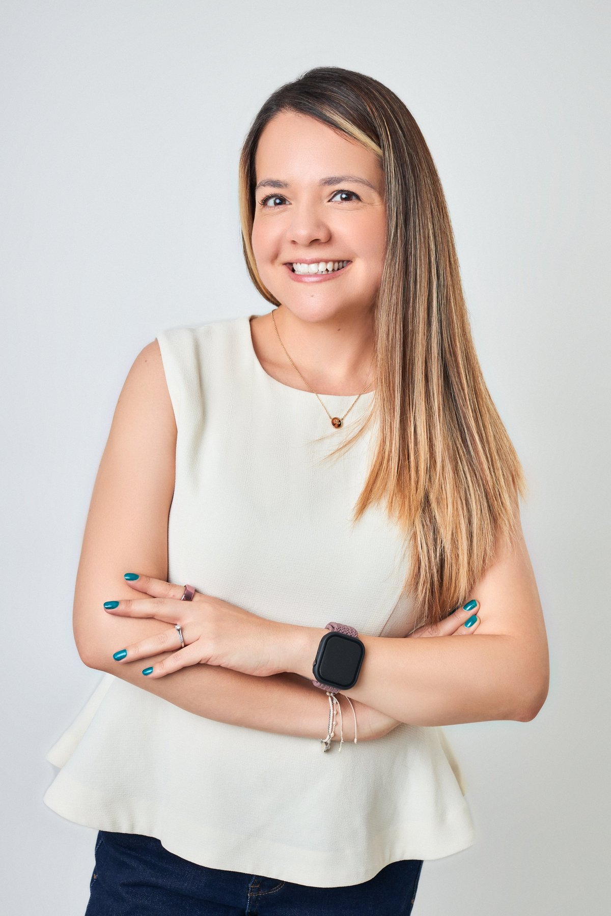 Smiling young woman with long brown hair wearing a cream sleeveless top, smartwatch, and blue nail polish against a white background