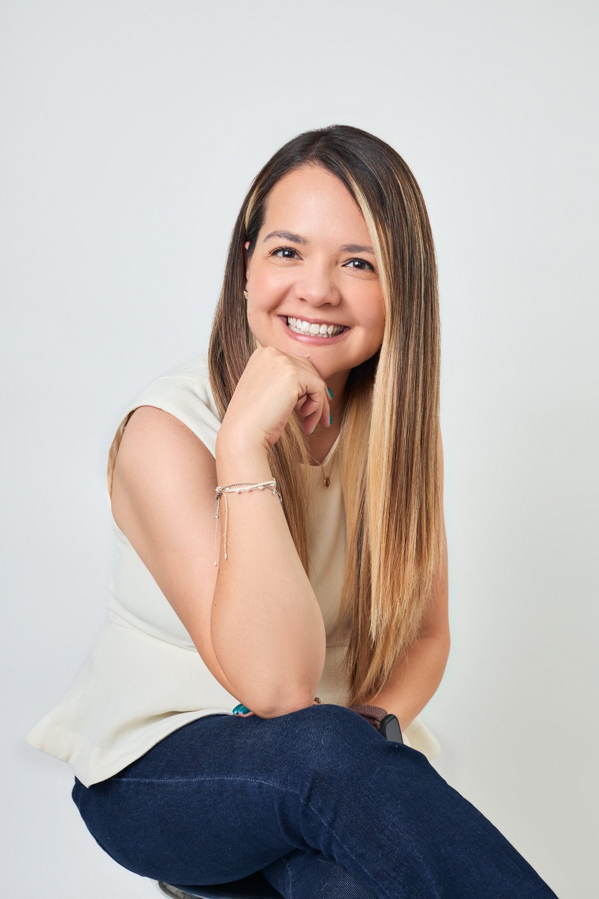 Young woman smiling with long brown hair, wearing a cream tank top and dark jeans, posing with hand near face